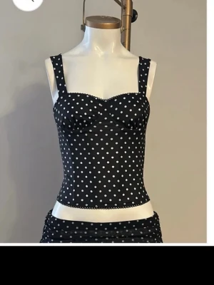Svart prickig  kosett topp - Svart croppad topp med vita prickar och breda axelband. Toppen har en söt, lätt markerad byst och fina spetsdetaljer längs kanterna. 
