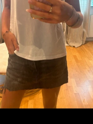 Svart jeanskjol med rå kant - Säljer en svart kort jeanskjol med rå, fransig kant och klassiska fem fickor. Kjolen har knapp och dragkedja framtill och är perfekt för en avslappnad och trendig look.