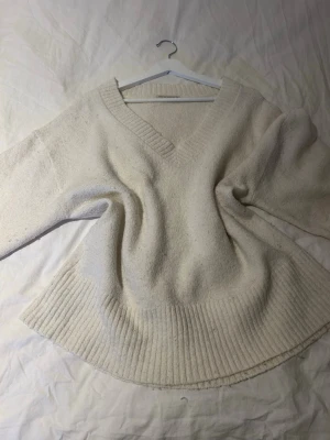 Krämvit stickad tröja med v-ringning - Mysig krämvit stickad tröja med djup v-ringning och bred ribbstickad kant nedtill. Tröjan har en avslappnad passform och långa ärmar, perfekt för lager på lager. Materialet känns mjukt och värmande, idealisk för kyligare dagar.