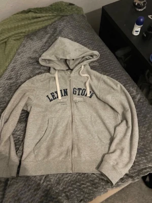 Grå hoodie från Lexington - Säljer en grå hoodie från Lexington med dragkedja och huva. Hoodien har vita snören, fickor framtill och Lexington-tryck i mörkblått på bröstet. Perfekt för chill dagar eller när du vill ha en enkel och snygg look.