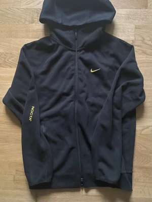 Nike nocta  - Svart hoodie från Nike NOCTA-kollektionen med hel dragkedja framtill och huva. Gula detaljer som Nike-logga på bröstet och NOCTA-tryck på ärmen. Tillverkad i mjukt fleecematerial, perfekt för en avslappnad streetwear-look.