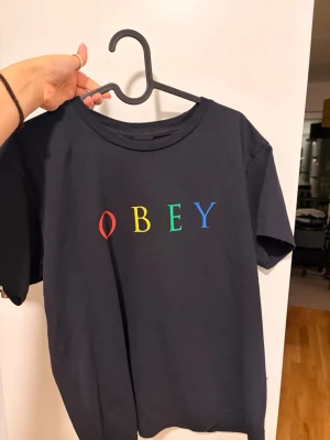 Svart OBEY t-shirt med färgglatt tryck - Säljer en svart t-shirt från OBEY med klassisk rund hals och korta ärmar. Framtill står OBEY i stora bokstäver i rött, gult, grönt och blått. Tillverkad i mjuk bomull för skön passform och enkel stil. Aldrig använd 