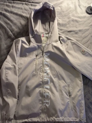 Beige vindjacka från Moncler - Snygg beige vindjacka från Moncler med huva och dragkedja framtill. Jackan har två sidofickor med dragkedja, en bröstficka och Moncler-logga på ärmen. Stora Moncler-bokstäver längs framsidan ger en exklusiv look. Tillverkad i lätt och tåligt material.