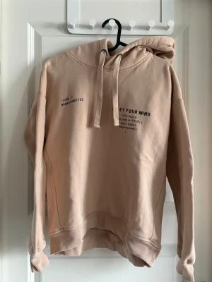 Beige hoodie med tryck - Snygg beige hoodie från lager 157 med svart texttryck på bröstet. Hoodien har en klassisk huva med dragsko och ribbade muddar vid ärmar och nederkant. Tillverkad i mjukt bomullsmaterial som känns skönt mot huden. Perfekt för en avslappnad streetwear-look.