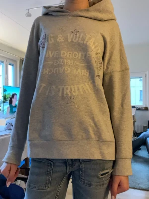 Grå hoodie Zadig & Voltaire - Snygg grå hoodie från Zadig & Voltaire med tryck och glittriga detaljer . Hoodien har en stor huva och avslappnad passform, perfekt till jeans. Tillverkad i mjukt sweatshirtmaterial för maximal komfort.