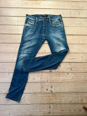 Blå jeans från Replay med slitningar - Säljer ett par blå jeans från Replay med snygga slitningar och tvättade detaljer. Jeansen har fem fickor, smal passform och klassisk knappgylf. Materialet är mjukt denim med stretch för extra komfort. Perfekta för en avslappnad och trendig look. Ett par defekter (se bilder)