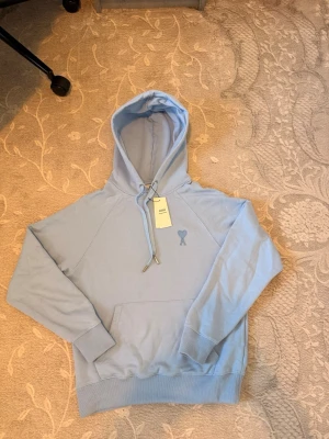 Ljusblå hoodie från Ami Paris - Säljer en ljusblå hoodie från Ami Paris med den ikoniska hjärtloggan broderad på bröstet. Hoodien har dragsko i huvan, magficka och ribbade muddar. Tillverkad i mjuk bomull för skön känsla och stilren look.
