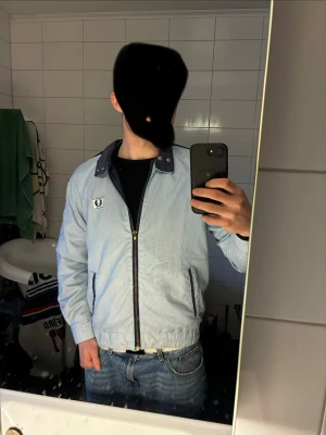 Ljusblå Fred Perry vindjacka - Snygg ljusblå vindjacka från Fred Perry med marinblå detaljer och dragkedja framtill. Jackan har ståkrage med tryckknappar, två fickor med dragkedja och elastisk midja. Ikonisk Fred Perry-logga på bröstet. Perfekt för dig som gillar retrostil.