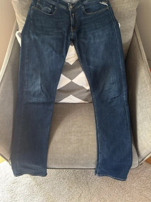 Replay Rocco 30/32 - Snygga mörkblå jeans från Replay med klassisk femficksmodell och raka ben. Jeansen har kontrastsömmar och detaljerade bakfickor med Replay-logga. Tillverkade i mjukt denimtyg som ger en skön passform och stilren look.