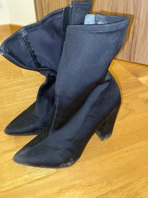 Svarta stretchiga stövletter med klack - Säljer ett par svarta stövletter med spetsig tå och hög, bred klack. Skorna är i ett stretchigt tygmaterial som sitter tight runt ankeln och har dragkedja på insidan. Från Nelly, storlek 38. Sulan torkas av innan jag skickar.