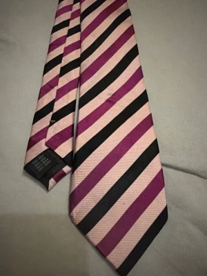 Randig slips i rosa och svart Liljewall design - Snygg slips med diagonala ränder i rosa, svart och lila. Tillverkad i ett glansigt tyg som ger en stilren look. Perfekt accessoar för att lyfta din outfit med lite färg och mönster.