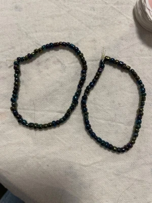 Två armband med mörka pärlor - Säljer två handgjorda armband med små, mörka pärlor i blå och lila toner. Armbanden har en enkel och stilren design med elastisk tråd som gör dem lätta att ta på och av. Perfekta för att addera en subtil färgklick till din outfit.OBS!!! Det finns bara två exemplar trots att det är tre på bilden. OBS! Materialet är okänt, kan ej garantera att det är rostfritt.