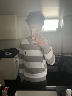 Randig tröja från Lyle & Scott - Snygg långärmad tröja från Lyle & Scott med breda beige och vita ränder. Tröjan har V-ringning och en gul broderad logga på bröstet. Ribbstickade muddar vid ärmslut och nederkant. Perfekt för en avslappnad och stilren look.