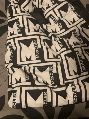 Moncler svartvit dunväst med logga - Säljer en fet svartvit dunväst från Moncler med stort grafiskt logomönster över hela jackan. Jackan har dragkedja och är fodrad med svart insida. Perfekt för dig som vill sticka ut i vinterkylan.