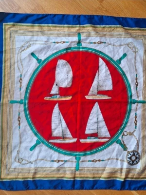 Färgglad sjal med segelbåtar, 80x80 cm TieRack - Säljer en snygg fyrkantig sjal i silkesliknande material, ca 80x80 cm. Sjalen har ett marint motiv med segelbåtar, röd mitt, blå kant och detaljer i grönt, guld och vitt. Perfekt accessoar för att lyfta din outfit med en nautisk touch.