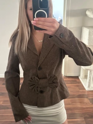 Brun kappa med rosett - Snygg brun kappa/kavaj med figurnära passform och rosettdetalj. Rosetten går att ta av eller flytta vars man vill. Mycket bra skick!