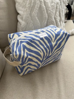 Blåvit necessär med zebramönster - Snygg necessär från Emelo i blått och vitt zebramönster. Tillverkad i bomull med praktiskt handtag på sidan och dragkedja upptill. Perfekt för att organisera smink eller småprylar i väskan. Stilren och trendig design som sticker ut. Vit insida med innerficka. Storlek large. Mått ca 27x17x12cm
