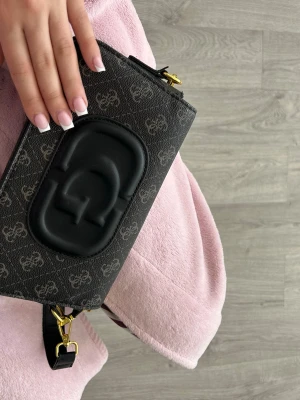 Svart axelremsväska från Guess - Snygg svart axelremsväska från Guess med GG-logga i relief framtill och diskret mönster över hela väskan. Väskan har guldfärgade metalldetaljer och justerbar axelrem. Perfekt för dig som vill ha en trendig och stilren handväska.