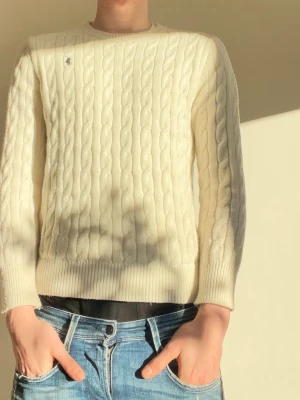 Vit kabelstickad tröja Polo Ralph Lauren - Kabelstickad tröja i mjuk vit bomull från Polo Ralph Lauren. Klassisk rund halsringning, ribbade muddar och nederkant samt den ikoniska lilla loggan broderad på bröstet. Perfekt till jeans för en clean och stilren look.