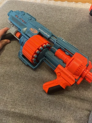 Nerf Elite 2.0 Shockwave blå/orange - Säljer en Nerf Elite 2.0 Shockwave blaster i blått och orange med roterande trumma för pilar. Den har detaljer i svart och grått och är tillverkad i plast. Perfekt för roliga Nerf-strider med vänner.