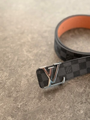 Svart rutigt LV-bälte i skinn - Snyggt svart bälte från Louis Vuitton med klassiskt rutigt mönster och stor LV-logga i silver på spännet. Bältet är gjort i skinn och har en lyxig känsla med orange insida. Perfekt accessoar för att lyfta din outfit.