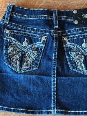 Mörkblå jeanskjol med broderade fickor - Säljer en mörkblå jeanskjol från Miss Me med snygga broderade detaljer på bakfickorna och kontrastsömmar. Kjolen har klassisk femficksdesign, dekorativa knappar och är i ett stretchigt denimtyg. Perfekt för dig som gillar en cool och avslappnad stil.