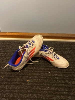 Vita Adidas fotbollsskor med röda detaljer - Säljer ett par vita Adidas fotbollsskor med röda och blå detaljer. Skorna har klassisk snörning, låg profil och är tillverkade i syntetmaterial för bra grepp och känsla på planen. Perfekta för dig som vill sticka ut på matchen! Tvättas innan frakten!
