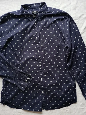 Mörkblå prickig skjorta från H&M - Snygg mörkblå skjorta från H&M med vita prickar. Skjortan har klassisk krage, lång ärm och knäppning framtill. Perfekt för dig som vill sticka ut lite extra med ett lekfullt mönster.