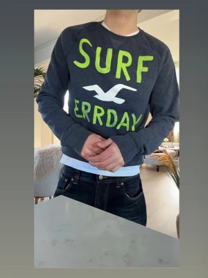 Mörkgrå sweatshirt från Hollister - Cool mörkgrå sweatshirt från Hollister med limegröna bokstäver och en vit mås på bröstet. Texten 'SURF ERRDAY' ger en avslappnad vibe. Tröjan har rund halsringning och långa ärmar, perfekt för en casual look. Storlek M. Jag är 184 på bilden! 
