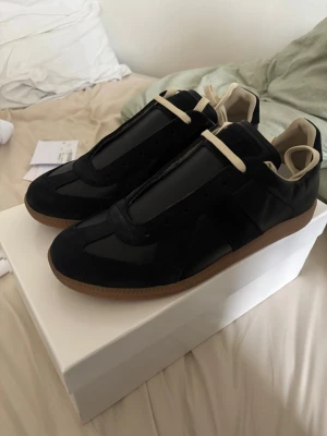 Svarta sneakers från Maison Margiela - Snygga svarta sneakers från Maison Margiela med beige detaljer och klassisk gummisula. Skorna har en mix av mocka och läder, beigea skosnören och en låg silhuett som ger en clean och stilren look.