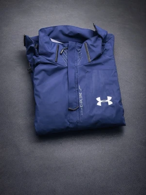 Under Armour  - Snygg blå regnjacka från Under Armour med huva och dragkedja framtill. Jackan har justerbara muddar och logga på bröstet. Tillverkad i vattenavvisande material, perfekt för regniga dagar. Stilsäker och funktionell design med sportig känsla.