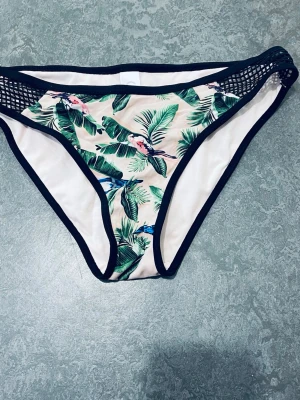 Blommiga bikiniunderdelar med meshdetalj - Snygga bikiniunderdelar med tropiskt blommönster i grönt och rosa, svarta kanter och meshdetaljer på sidorna. Perfekta för sommaren och strandhäng. Bekväm passform och trendig design.