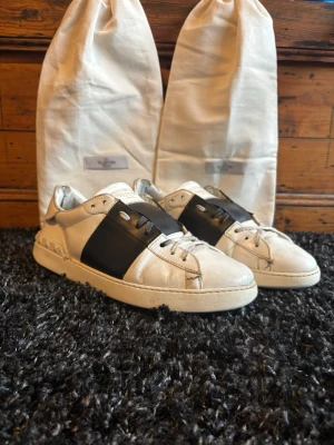 Valentino Open vita sneakers med svart detalj - Snygga vita sneakers från Valentino med bred svart läderdetalj över snörningen. Klassisk låg modell med rund tå och vita skosnören. Tillverkade i läder och kommer med original dustbags. Perfekta för dig som gillar exklusiv och stilren design. Skorna är lite slitna som ni ser och det är en extra sula över originalet för den har tappat mycket färg, sen är ett skosnöre trasigt men knutet innanför läderlappen i mitten av skon så det syns inte. Pågrund av de här är priset hyfsat lågt, kvitto finns!!!