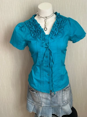 Turkos blus med volangkrage - Snygg turkos blus med korta puffärmar och volangdetaljer runt kragen. Knäppning framtill och dekorativa sömmar längs bröstet. Tillverkad i ett lätt bomullsliknande material som ger en fräsch och ungdomlig look.