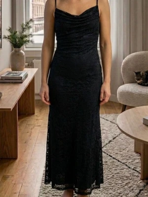 Svart långklänning med spetsdetaljer - Elegant svart långklänning med tunna axelband och vacker spets över hela klänningen. Klänningen har en smickrande passform och lätt draperad urringning. Perfekt för dig som vill ha en stilren och tidlös look.