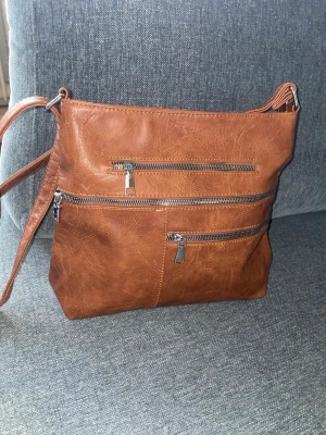 Brun axelväska i konstläder - Snygg brun axelväska i fejkläder med justerbar axelrem och flera dragkedjeförsedda fack både fram och bak. Perfekt för dig som vill ha ordning på dina grejer och gillar en stilren look. Väskan har silverfärgade detaljer och en mjuk. Funktionella dragkedjor framtill som kan öppnas och ger praktiska extra fack. Marknadsköpt väska utan specifikt märke 🌸