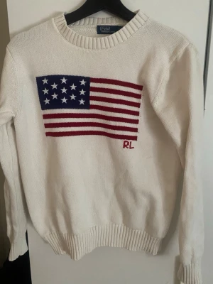 Vit stickad tröja Polo Ralph Lauren - Säljer en vit stickad tröja från Polo Ralph Lauren med amerikansk flagga i blått, rött och vitt på bröstet samt RL-broderi. Tröjan har rund halsringning, långa ärmar och ribbade muddar. Perfekt för dig som gillar klassisk preppy stil. Är inte äkta och är lite för små, så den funkar också som XS