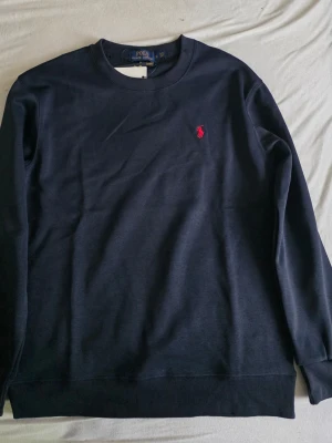 Fleece Crewneck Sweatshirt - Klassisk mörkblå sweatshirt från Polo Ralph Lauren med rund halsringning och röd broderad logga på bröstet. Tröjan har ribbade muddar vid ärmslut och nederkant.