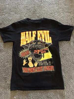 VINTAGE T SHIRT  - Svart t-shirt från HALF EVIL med stor grafisk print framtill i gult, rött och grått. Trycket har en varg, texten 'HALF EVIL' och retro skräckfilmskänsla. Klassisk passform och rund hals. Perfekt för dig som gillar streetwear och statement-plagg.