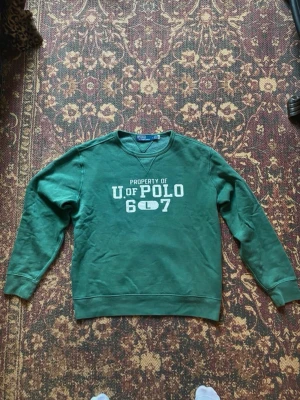 Grön sweatshirt från Polo Ralph Lauren - Grön sweatshirt med trycket 'PROPERTY OF U. OF POLO 607' på bröstet. Klassisk crewneck-modell med ribbade muddar vid ärmslut och nederkant. Perfekt för en avslappnad och sportig stil.
