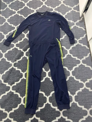 Blått träningsset från Lacoste - Snyggt marinblått träningsset från Lacoste med matchande tröja och byxor. Setet har limegröna och vita detaljer längs ärmar och ben, samt den klassiska Lacoste-loggan på bröstet och benet. Tillverkat i ett lätt och sportigt material, perfekt för träning eller chill.