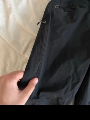 Svarta cargobyxor med dragkedjor, haglövs - Säljer ett par svarta cargobyxor med dragkedjeförsedda fickor och justerbara benslut med dragsko. Byxorna har en clean look och är tillverkade i ett lätt, slitstarkt material som passar perfekt för en aktiv vardag., Haglöfs.