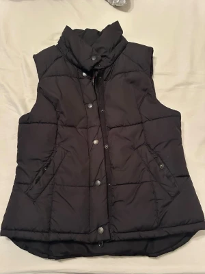 Svart quiltad dunväst med hög krage - Snygg svart dunväst med hög krage och tryckknappar framtill. Västen har två fickor med knappstängning och är quiltad för extra värme. Perfekt för lager på lager och en clean look.