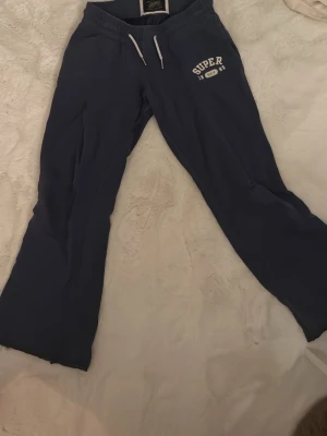 Mörkblå mjukisbyxor Superdry - Mörkblå mjukisbyxor från Superdry med vita dragsnören i midjan och tryckt logga på vänster sida. Byxorna har raka ben och resår i midjan för extra komfort. Perfekta för chill dagar eller när du vill vara bekväm. ( Jag säljer även matchande hoodie till så är du intresserad så kan jag även skicka bild på den).☺️( Jag säljer byxorna för 250kr, hoodie för 300kr och hela settet tillsammans för 500kr).