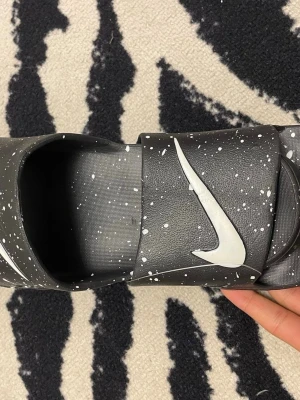 Nike Vapormax svarta sandaler - Svarta Nike Vapormax sandaler med vitt stänkmönster och stor vit Swoosh-logga på ovandelen. Sandalerna har en chunky sula och texten 'Vapormax' på sidan. Tillverkade i syntetmaterial för en sportig och modern look.