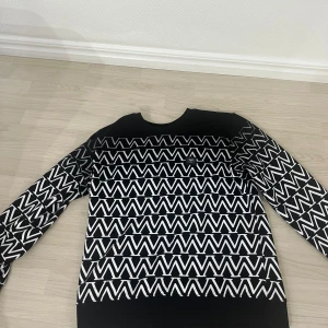 Svartvit tröja med zigzag-mönster - Snygg svart tröja med vitt zigzag-mönster över hela plagget. Tröjan har rund halsringning och långa ärmar. Materialet ser ut att vara mjukt och stretchigt, perfekt för en trendig och avslappnad stil.