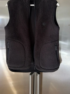 Svart fleece j Lindberg väst  - Mysig svart teddyväst med hög krage och hel dragkedja framtill. Västen har två stora fickor med dragkedja och en liten logodetalj på bröstet. Perfekt för lager-på-lager och ger en cool, avslappnad look.