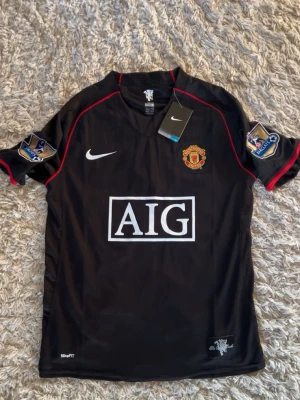 Manchester United Ronaldo tröja svart - Säljer en svart Manchester United fotbollströja från Nike med röda detaljer och AIG som sponsor. Tröjan har Premier League-patchar på ärmarna och Ronaldo 7 tryckt på ryggen. Perfekt för dig som älskar fotboll och vill ha en ikonisk supportertröja.