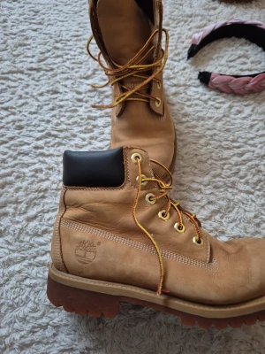 Beige Timberland kängor i läder - Klassiska beige Timberland kängor i läder med robust sula och gulbruna snören. Kängorna har svart vadderad kant upptill och metallfärgade öljetter. Perfekta för dig som vill ha en ikonisk streetwear-look.