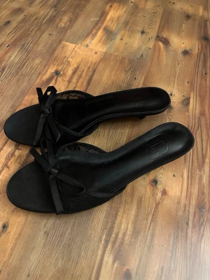 Svarta pumps med rosett och spetsdetalj - Säljer ett par svarta pumps med låg klack, öppen tå och fina rosetter framtill. Skorna har spetsdetaljer på sidorna som ger en elegant touch. 3cm klack, aldrig använda 